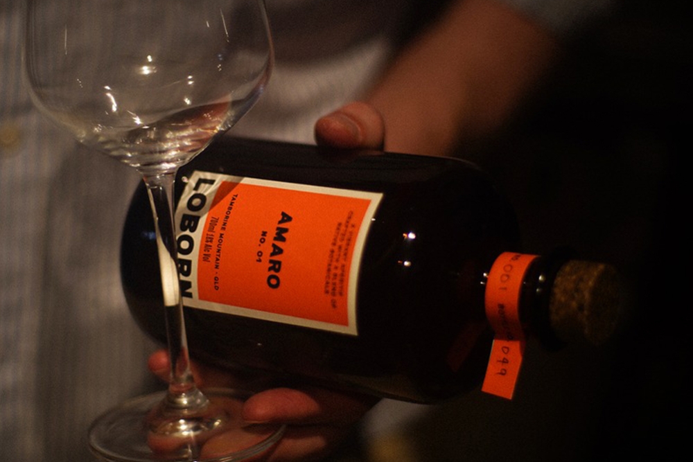 A hand holding a bottle of amaro on its side with an empty glass on a table at Loborn Tamborine Mountain 